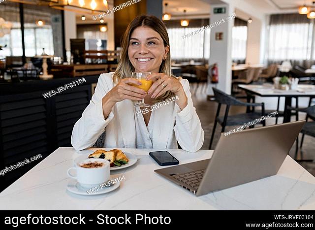 Smiling businesswoman holding juice glass sitting with laptop at restaurant