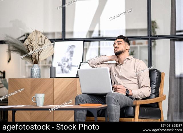 Short break. Young bearded man with closed eyes touching his neck sitting with laptop in armchair having rest