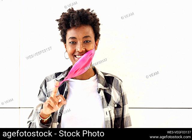 Smiling young woman removing pink mask against white wall