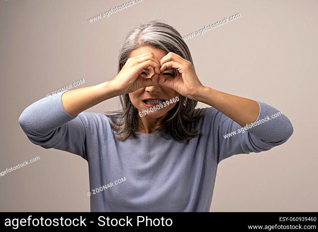 Irritated Woman Grits Her Teeth While Gesturing Glasses With Her Hands. Angry Grey-Haired Woman Looking Through Her Hands As Through Binoculars And Gritting Her...