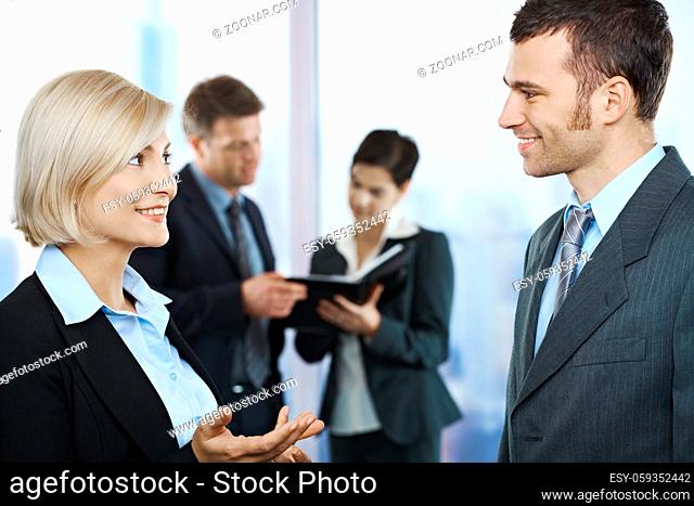 Standing business people talking in office smiling at each other, coworkers looking at documents in background