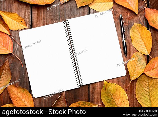 open notebook with blank white sheets and yellow cherry leaves on brown wooden background, top view