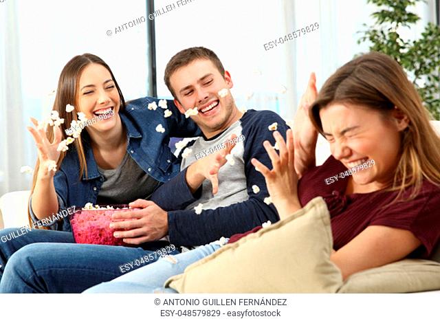 Three happy friends joking and throwing popcorn sitting on a sofa in the living room at home