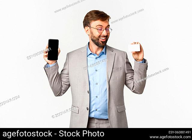 Cheerful male entrepreneur with beard, wearing grey suit and glasses, showing app on mobile phone screen and credit card, smiling excited