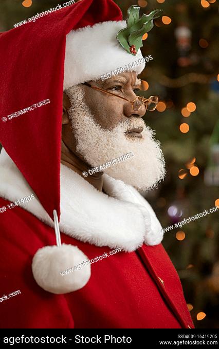 An African American man dressed as Santa Claus sitting in front of a Christmas tree turned profile, looking off camera