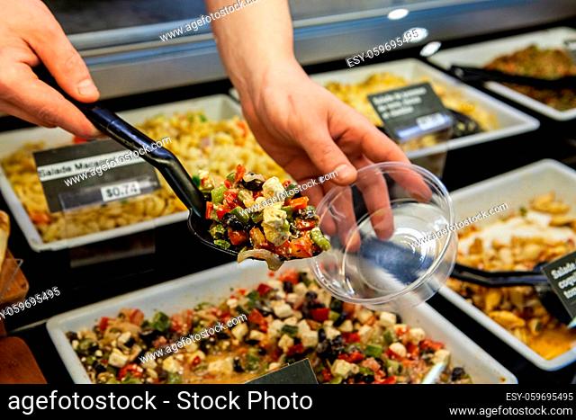 A person is viewed closeup, filling a small plastic tub with prepared greek feta salad from a delicatessen counter in a food hall