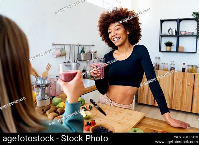Happy young woman holding glass of smoothie talking with friend in kitchen