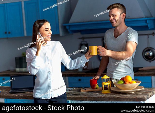 Brunette woman talking on phone whille her husband giving her cup of tea. Couple in the morning in kitchen, woman is ready to go to work while husband stays at...