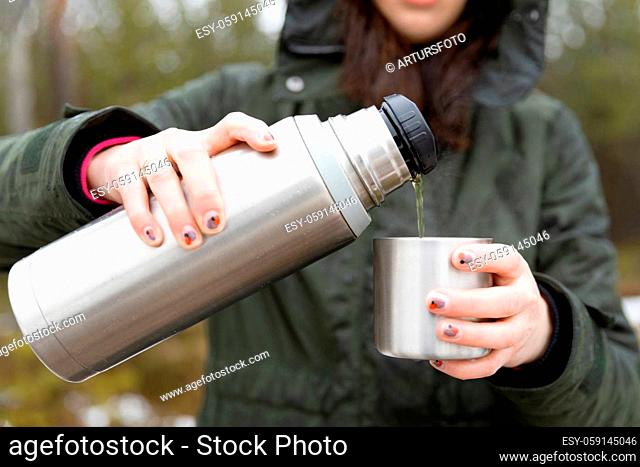 Youg beautifull women pouring beverage from termos to cup in a cold raini day. Healthy living and hiking