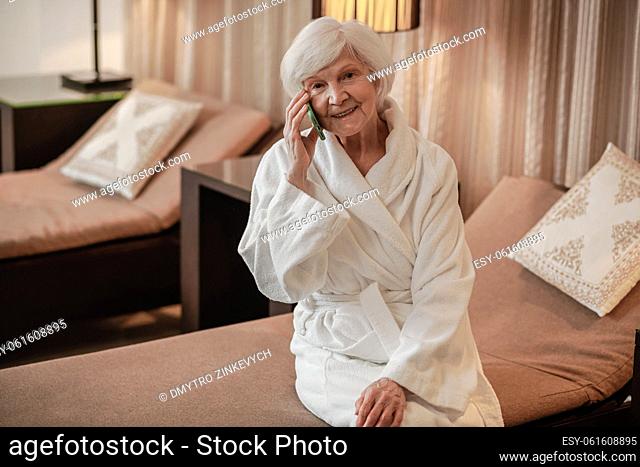 Advanced senior. Good-looking elderly woman in white robe with a phone in hands
