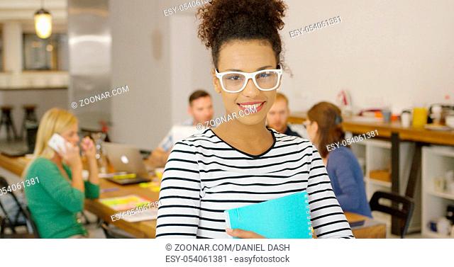 Beautiful ethnic girl in glasses holding notepad and smiling cheerfully at camera on background of office table with coworkers