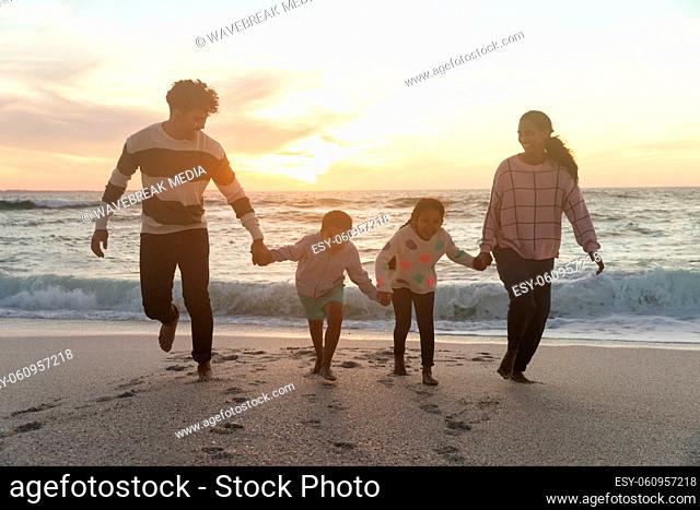 Full length of happy multiracial family holding hands running at beach against sky during sunset