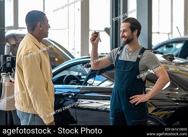 Gas station. Smiling caucasian bearded worker with car key looking confidently at dark-skinned customer standing in gas station