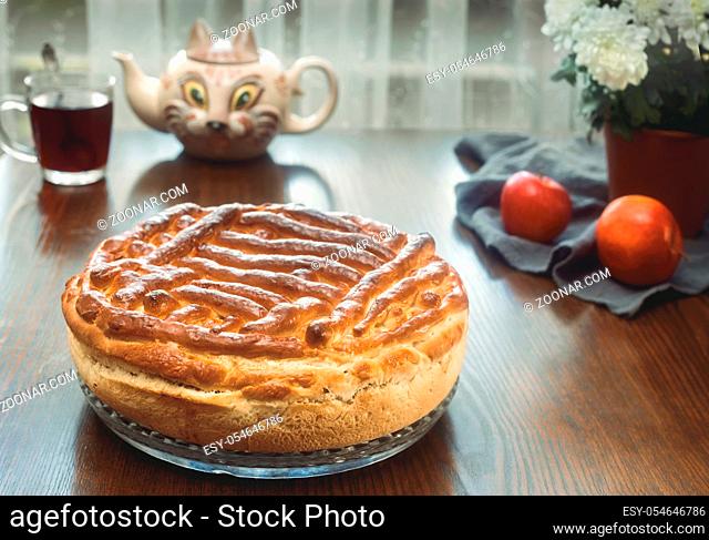 On the dining table on a glass tray is a delicious large homemade cake. On a napkin near tea in a glass and apples