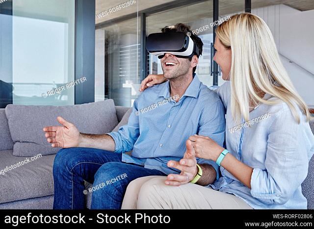 Blond woman with arm around looking at cheerful man wearing virtual reality simulator in living room