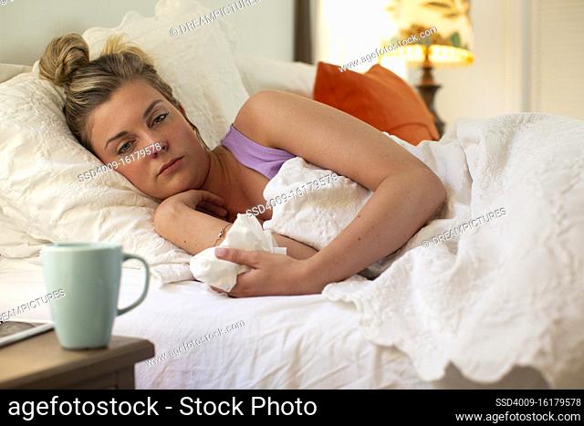 Young woman in her bed debating getting out of bed, holding tissue in hand, smartphone and coffee mug sitting on bedside table
