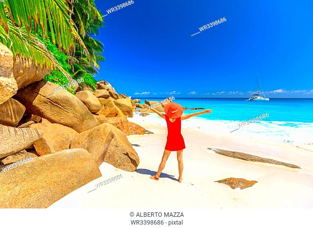 Tourist woman in red dress looking at turquoise sea and sailing boat and catamarans on horizon, Praslin Island, Seychelles, Indian Ocean, Africa