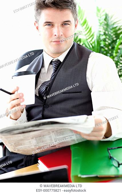 Serious businessman drinking a coffee while reading a newspaper sitting at his desk