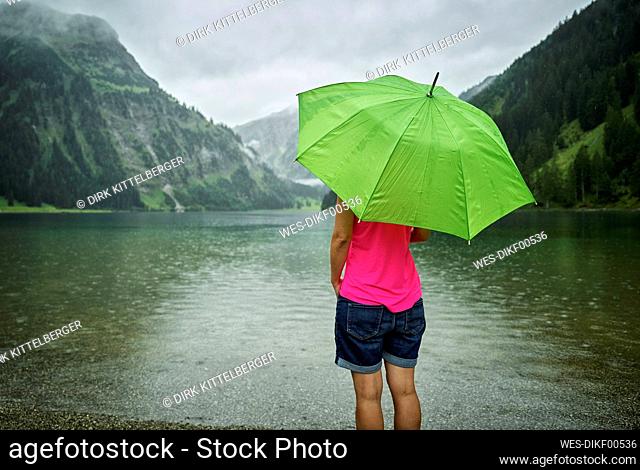 Mature woman standing with green umbrella at Vilsalpsee lakeshore during rainy season