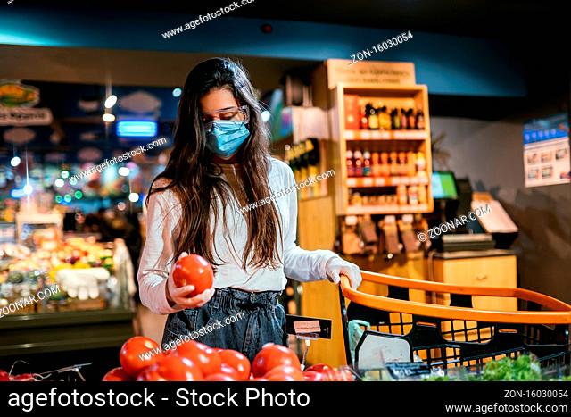 Woman with the surgical mask and the gloves is shopping in the supermarket after coronavirus pandemic. The girl with surgical mask is going to buy tomatos