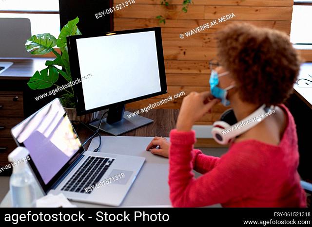 Mixed race woman wearing face mask using computer at desk in casual office