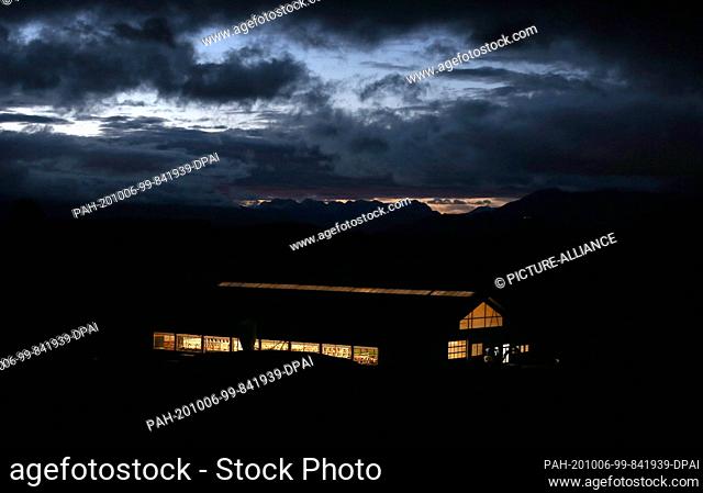dpatop - 06 October 2020, Bavaria, Oy-Mittelberg: Cows stand in the stable of a farm just before sunrise in front of the cloudy Alps