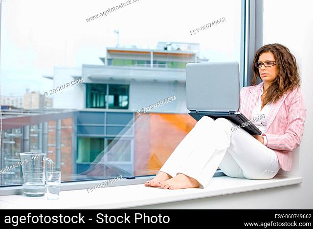 Young creative businesswoman sitting at office window and working on laptop computer