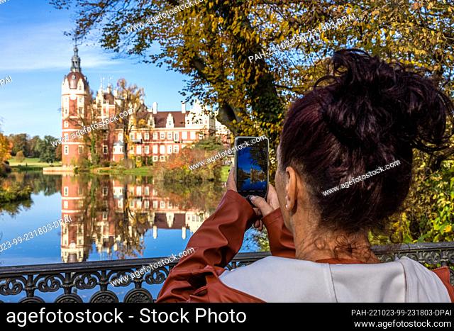 23 October 2022, Saxony, Bad Muskau: A young woman takes a photo of the New Castle reflected in a pond in Prince Pückler Park with her smartphone in the...