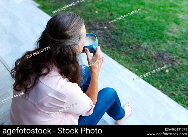 Happy senior caucasian woman sitting on stairs, drinking coffee in garden