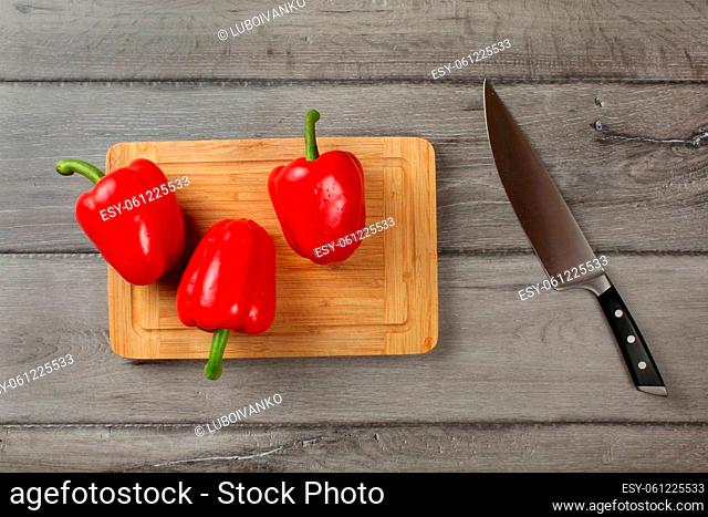 Tabletop view, three red bell peppers, sprayed with drops of water, ready to be cut on chopping board, with chef knife at side