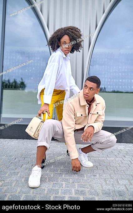 Style. Stylish confident dark-skinned guy crouched and girl in glasses with bag posing on city street near building