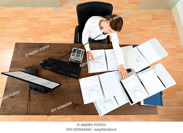 High Angle View Of Young Businesswoman Attending Call While Calculating Finance At Desk