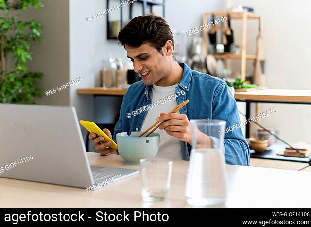 Smiling man using mobile phone while having food at home