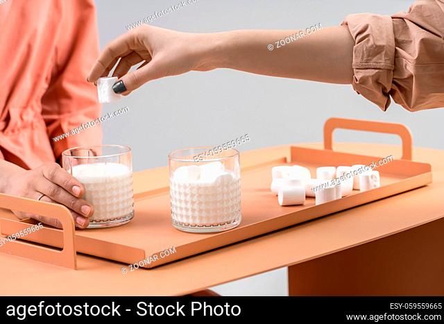 Two glasses with milk and marshmallows on the orange metal tray on the same table on a gray background in a studio. Couple of girls sitting near the table