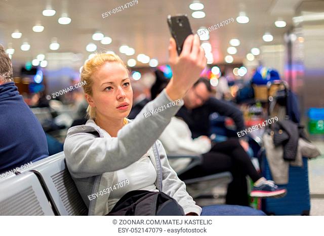 Casual blond young woman taking selfie photo using her cell phone while waiting to board a plane at the departure gates