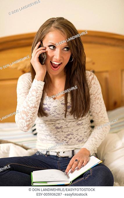 Teenage girl lying on a bed talking on a cell phone while doing homework