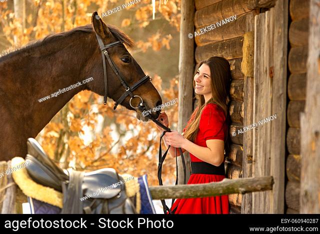 Portrait of a beautiful girl in a red dress, a girl holding a horse by the bridle, a beautiful background of a wooden wall and forest