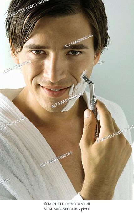 Man in bathrobe shaving, smiling at camera, close-up