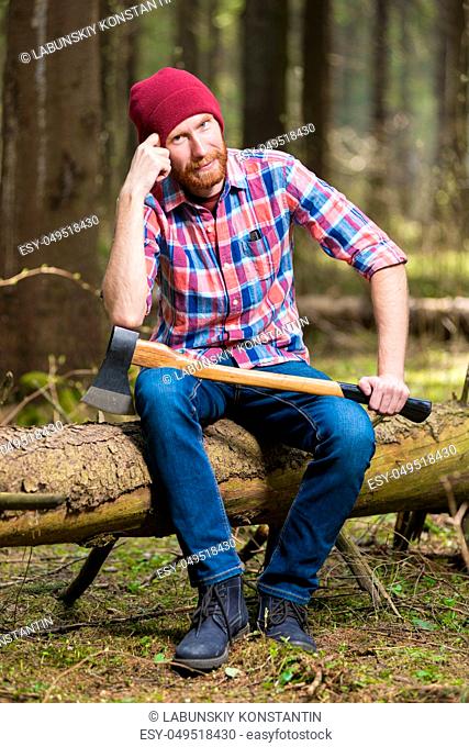 thoughtful lumberjack in a hat and shirt with an ax sits on a log in the woods