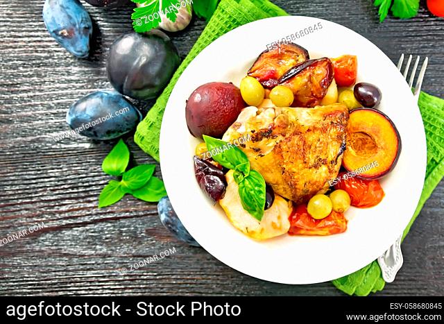 Baked chicken with tomatoes, apples, plums and grapes in a plate on a napkin, garlic, parsley and basil on wooden board background from above
