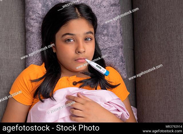 Close-up portrait of a sick girl lying on sofa with a digital thermometer on her mouth