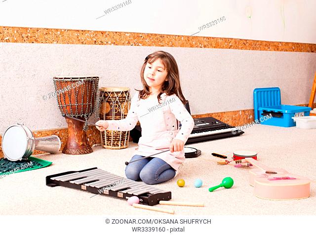 The cute little girl sits on the floor and playing on the xylophone at the kindergarten. The girl is testing various musical instruments