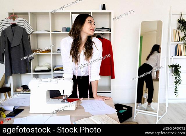 Female fashion designer looking away while leaning on desk in studio