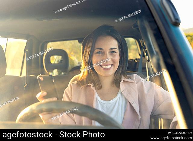Smiling young woman holding mobile phone in car during sunset