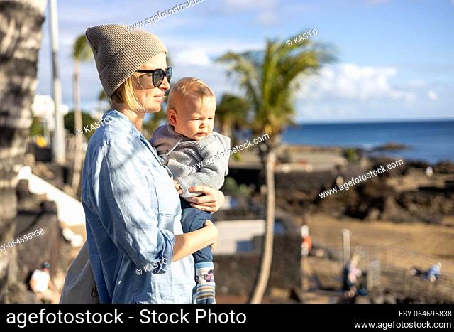Fashinable mother holding her infant boy child in lap enjoing panoramic view along palm lined beach promenade of Puerto del Carmen, Lanzarote, Canary islands