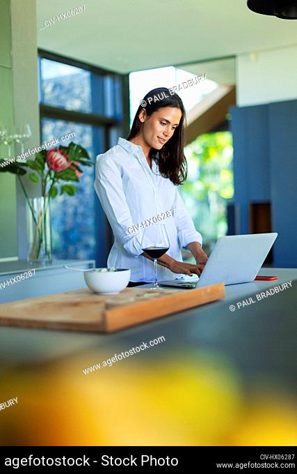 Woman using laptop and drinking red wine in kitchen