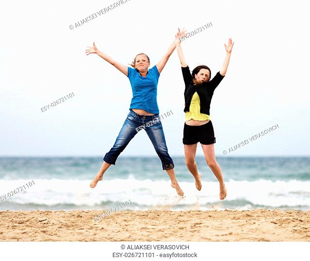 Happy girls jumping outdoors. Two happy young women jump on the beach against the sea and cloudless sky. Selective focus on the models