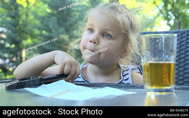 Cute little girl sits thoughtfully at the table with a lens in her hand. Close-up of blonde girl holding a magnifying glass in her hand and thinking about...