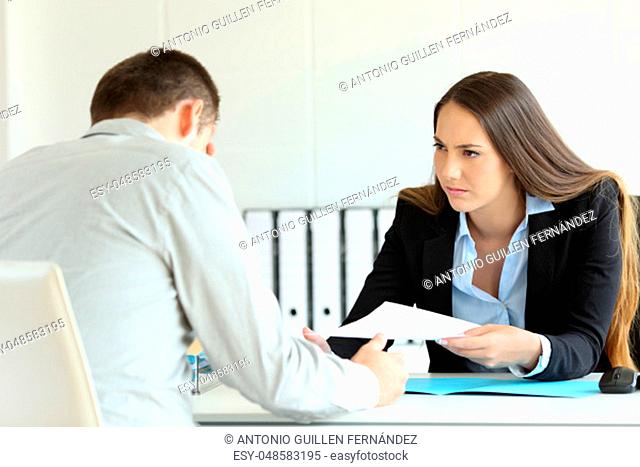 Angry boss giving a document to a sad employee sitting in a desk at office