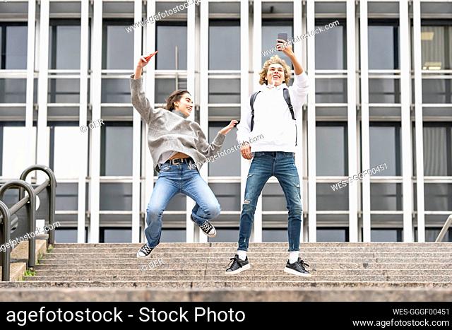 Carefree friends with taking selfie through smart phone while jumping over steps against building